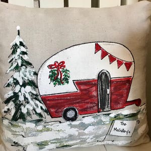 Puede incluir: Una almohada beige con un dise&ntilde;o pintado a mano de una caravana roja y blanca con una corona de Navidad en el lateral. La caravana tiene una puerta negra y una rueda negra. Hay un pino verde con nieve blanca en las ramas en el fondo. El texto "The McCulley's" est&aacute; escrito en un cartel blanco en la esquina inferior derecha.