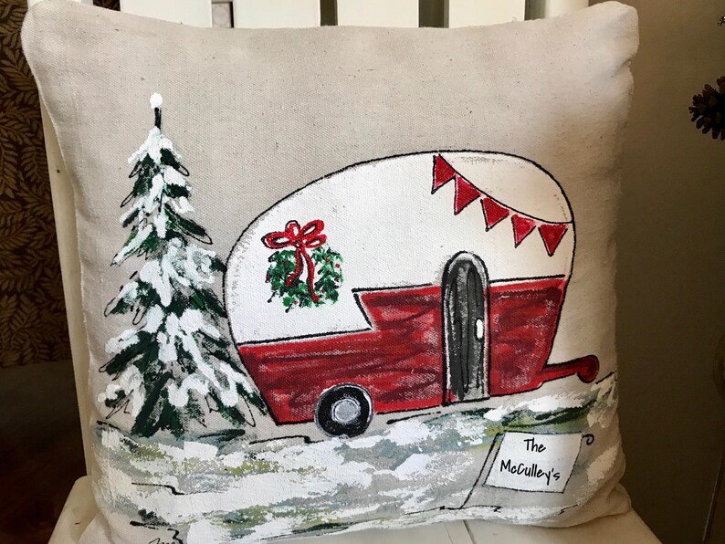 Puede incluir: Una almohada beige con un dise&ntilde;o pintado a mano de una caravana roja y blanca con una corona de Navidad en el lateral. La caravana tiene una puerta negra y una rueda negra. Hay un pino verde con nieve blanca en las ramas en el fondo. El texto "The McCulley's" est&aacute; escrito en un cartel blanco en la esquina inferior derecha.