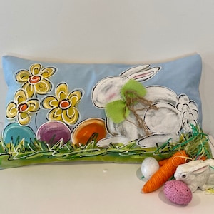 May include: A blue and white pillow with a painted design of a white bunny, yellow flowers, and colorful Easter eggs. The bunny is wearing a green bow and is surrounded by green grass.