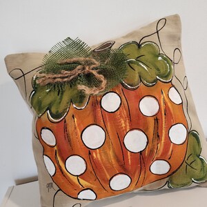 Puede incluir: Una almohada beige con una calabaza naranja pintada a mano con lunares blancos. La calabaza tiene hojas verdes y un lazo de arpillera marr&oacute;n.