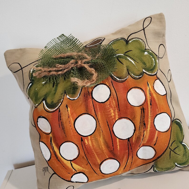 Puede incluir: Una almohada beige con una calabaza naranja pintada a mano con lunares blancos. La calabaza tiene hojas verdes y un lazo de arpillera marr&oacute;n.