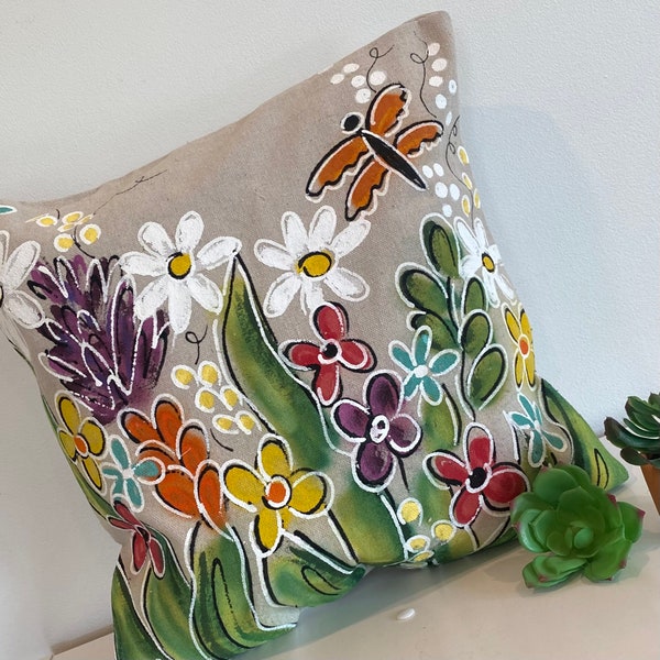 May include: A beige linen pillow with a hand-painted design of colorful flowers and a butterfly. The flowers are in shades of white, red, orange, yellow, purple, and green.