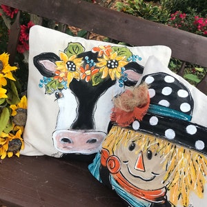 May include: Two decorative throw pillows with painted designs. One pillow features a black and white cow with a sunflower and blue flower crown. The other pillow features a smiling scarecrow with a black and white polka dot hat and a brown, orange, and blue scarf.