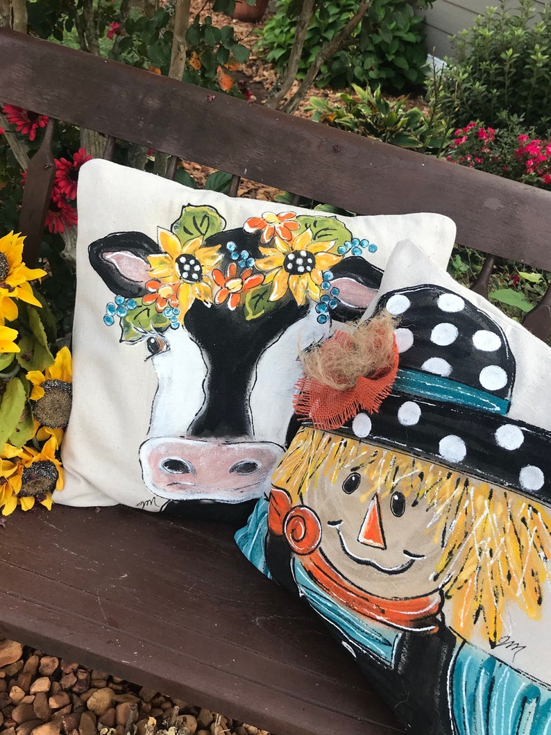 May include: Two decorative throw pillows with painted designs. One pillow features a black and white cow with a sunflower and blue flower crown. The other pillow features a smiling scarecrow with a black and white polka dot hat and a brown, orange, and blue scarf.
