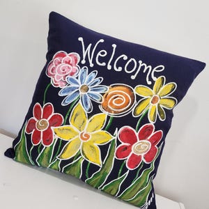 May include: A navy blue throw pillow with a hand-painted design of colorful flowers and the word "Welcome".