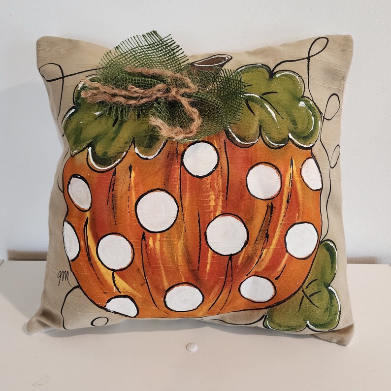 Puede incluir: Una almohada beige con una calabaza naranja pintada a mano con lunares blancos. La calabaza tiene hojas verdes y un lazo de arpillera marr&oacute;n.
