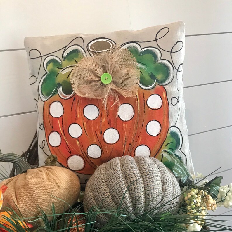 May include: A decorative pillow with a hand-painted orange pumpkin with white polka dots and green leaves. The pumpkin has a burlap bow with a green button in the center. The pillow is on a white background.