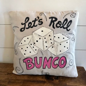Peut inclure: Un coussin carr&eacute; gris clair avec les mots "Let's Roll BUNCO" en noir et rose. Trois d&eacute;s blancs avec des points noirs sont au centre. Le coussin est pos&eacute; sur une surface en bois.