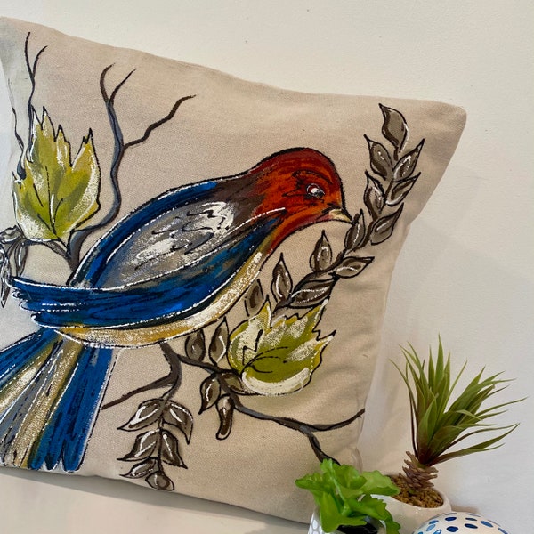 May include: A beige throw pillow with a hand-painted design of a blue bird with red and yellow accents perched on a branch with green leaves.