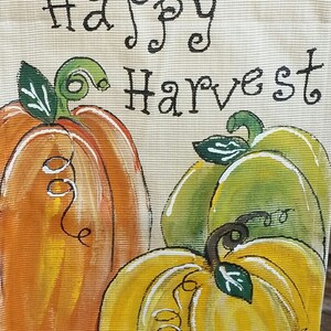 May include: A hand-painted burlap banner with the text "Happy Harvest" and three pumpkins. The pumpkins are painted in shades of orange, yellow, and green.