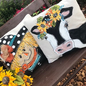 May include: Two decorative throw pillows with painted designs. One pillow features a scarecrow with a black and white polka dot hat and a brown bow. The other pillow features a black and white cow with a floral crown.