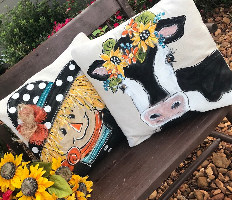 May include: Two decorative throw pillows with painted designs. One pillow features a scarecrow with a black and white polka dot hat and a brown bow. The other pillow features a black and white cow with a floral crown.