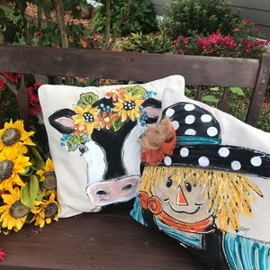 May include: Two decorative throw pillows on a wooden bench. One pillow features a black and white cow with a sunflower crown. The other pillow features a scarecrow with a black and white polka dot hat.