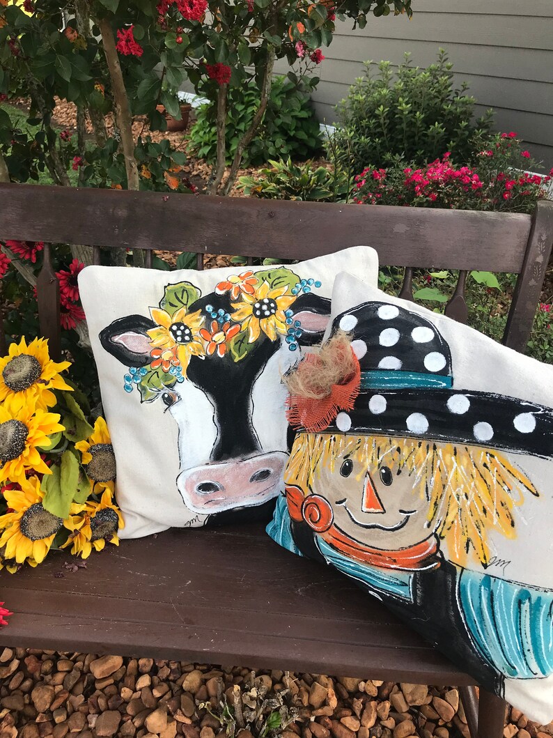 May include: Two decorative throw pillows on a wooden bench. One pillow features a black and white cow with a sunflower crown. The other pillow features a scarecrow with a black and white polka dot hat.