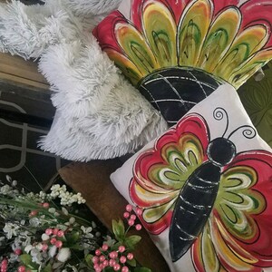 K&ouml;nnte beinhalten: Zwei dekorative Kissen mit handgemalten Designs. Ein Kissen zeigt einen roten, gelben und gr&uuml;nen Schmetterling mit schwarzen Akzenten. Das andere Kissen zeigt eine rote, gelbe und gr&uuml;ne Blume mit schwarzen Akzenten.