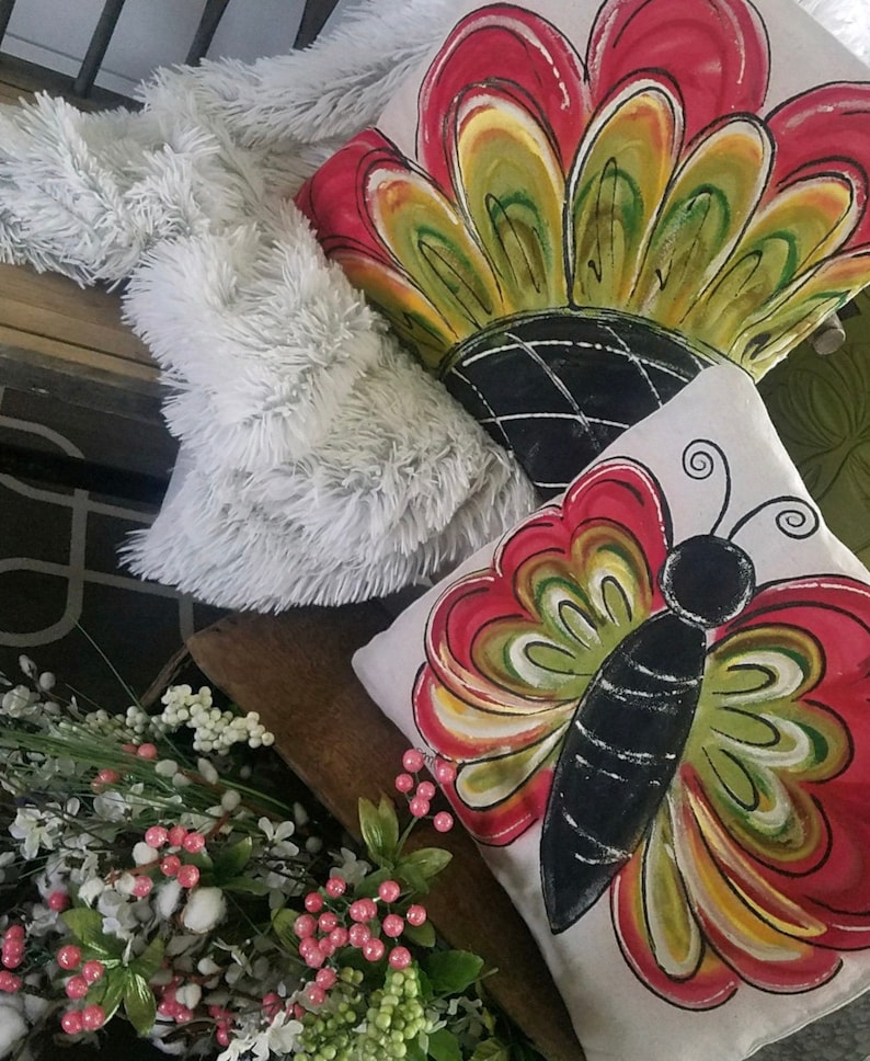 K&ouml;nnte beinhalten: Zwei dekorative Kissen mit handgemalten Designs. Ein Kissen zeigt einen roten, gelben und gr&uuml;nen Schmetterling mit schwarzen Akzenten. Das andere Kissen zeigt eine rote, gelbe und gr&uuml;ne Blume mit schwarzen Akzenten.