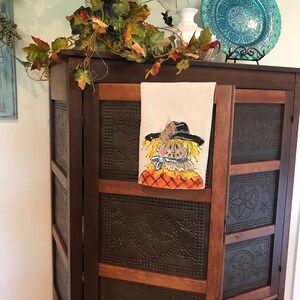 May include: A brown wooden cabinet with a distressed finish and a single drawer. The cabinet has a tin panel with a decorative pattern and a white towel with a painted scarecrow design hanging on the door.