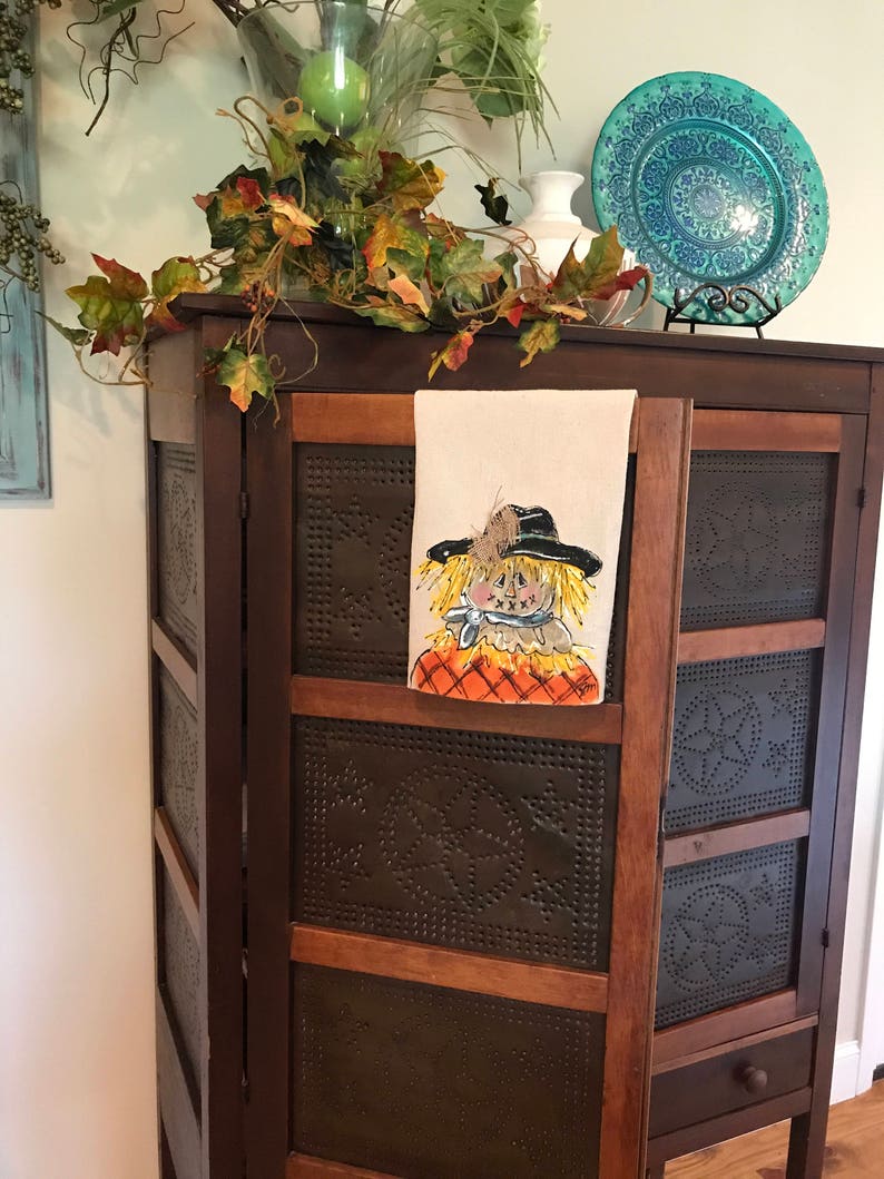 May include: A brown wooden cabinet with a distressed finish and a single drawer. The cabinet has a tin panel with a decorative pattern and a white towel with a painted scarecrow design hanging on the door.