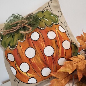 Puede incluir: Una almohada beige con una calabaza naranja pintada a mano con lunares blancos. La calabaza tiene un tallo y hojas verdes. La almohada est&aacute; decorada con un lazo de arpillera marr&oacute;n y hojas de arpillera verdes.