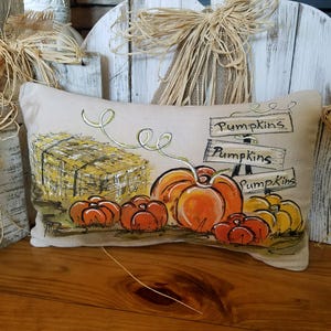 May include: A beige pillow with a painted design of pumpkins, hay bales, and a sign that reads "Pumpkins".