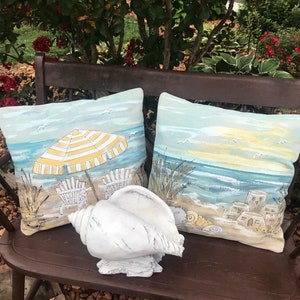 May include: Two decorative cushions featuring beach scenes, one with a yellow and white striped umbrella and beach chairs, the other with a sunset over the sea. A white seashell sits in front of the cushions on a brown wooden bench.