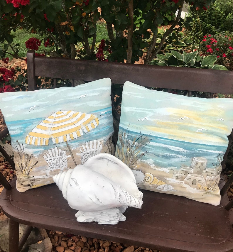 May include: Two decorative cushions featuring beach scenes, one with a yellow and white striped umbrella and beach chairs, the other with a sunset over the sea. A white seashell sits in front of the cushions on a brown wooden bench.