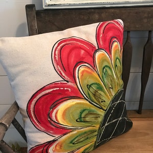 K&ouml;nnte beinhalten: Ein beiges Kissen mit einem gro&szlig;en, handgemalten Blumendesign. Die Blume ist rot, gelb, gr&uuml;n und schwarz. Das Kissen liegt auf einer Holzbank.