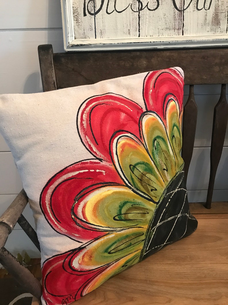 K&ouml;nnte beinhalten: Ein beiges Kissen mit einem gro&szlig;en, handgemalten Blumendesign. Die Blume ist rot, gelb, gr&uuml;n und schwarz. Das Kissen liegt auf einer Holzbank.