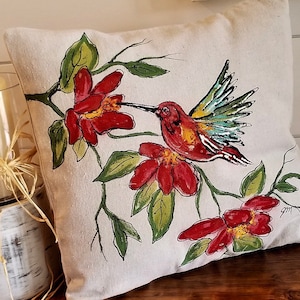 May include: A white pillow with a hand-painted design of a hummingbird in flight, feeding from a red flower. The hummingbird is painted in shades of red, yellow, green, and blue. The flowers are painted in shades of red and green. The pillow is a great addition to any home decor.