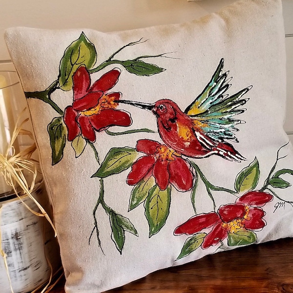 May include: A white pillow with a hand-painted design of a hummingbird in flight, feeding from a red flower. The hummingbird is painted in shades of red, yellow, green, and blue. The flowers are painted in shades of red and green. The pillow is a great addition to any home decor.