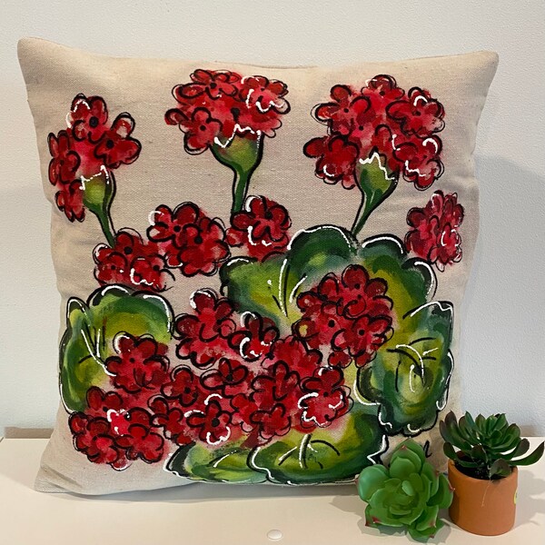 May include: A decorative throw pillow with a hand-painted design of red geraniums and green leaves on a beige linen fabric.