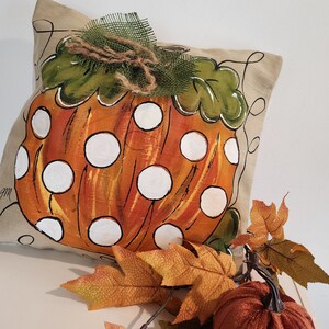 Puede incluir: Una almohada beige con una calabaza naranja pintada a mano con lunares blancos. La calabaza tiene un tallo y hojas verdes. La almohada est&aacute; decorada con un lazo de arpillera marr&oacute;n y una malla verde.