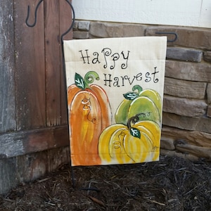 May include: A beige garden flag with the text "Happy Harvest" in black cursive lettering. Three painted pumpkins are featured on the flag: one orange, one yellow, and one green.