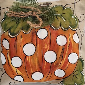 Puede incluir: Una almohada decorativa con una calabaza naranja pintada a mano con lunares blancos y hojas verdes. La calabaza est&aacute; rodeada de un lazo de arpillera.