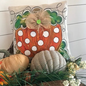 May include: A decorative pillow with a hand-painted orange pumpkin with white polka dots. The pumpkin has a green stem and a burlap bow with a green button. The pillow is on a white background.