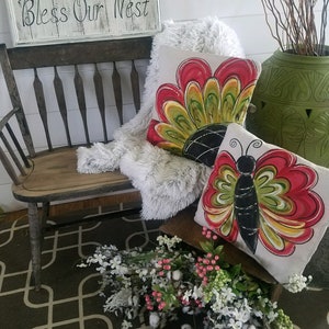 K&ouml;nnte beinhalten: Zwei dekorative Kissen mit einem Schmetterlings- und Blumendesign. Die Kissen liegen auf einer Holzbank mit einem Schild, auf dem "Bless Our Nest" steht.