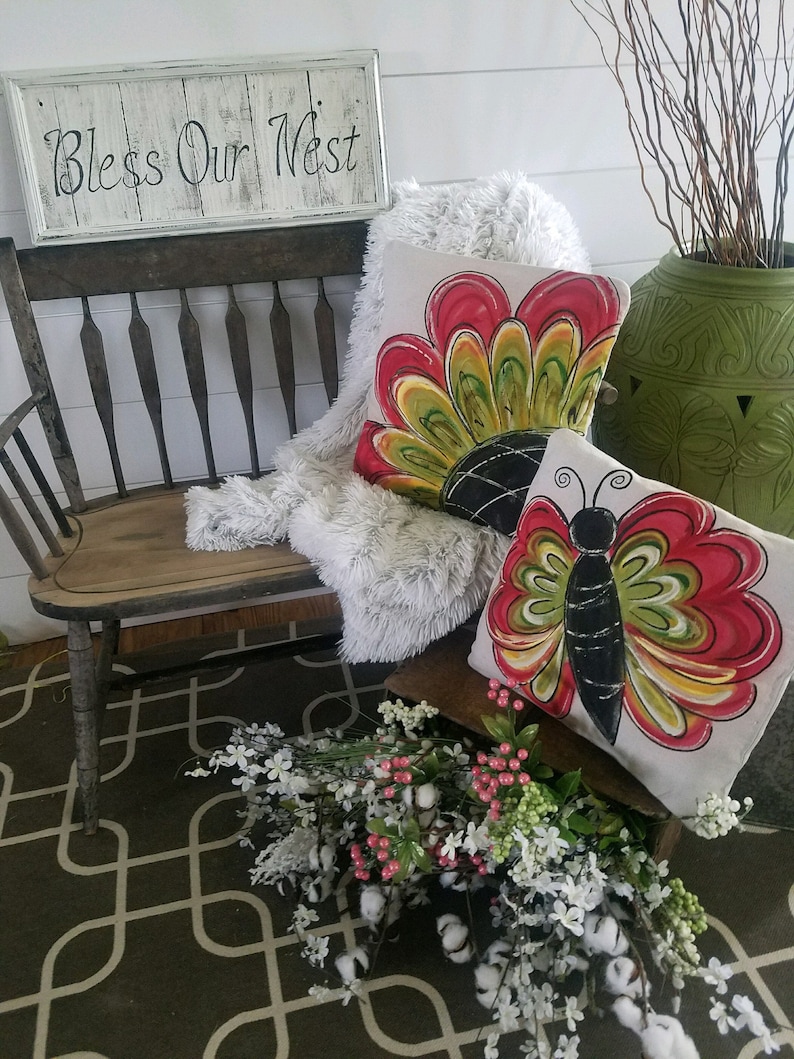 K&ouml;nnte beinhalten: Zwei dekorative Kissen mit einem Schmetterlings- und Blumendesign. Die Kissen liegen auf einer Holzbank mit einem Schild, auf dem "Bless Our Nest" steht.