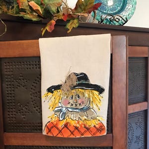 May include: A hand-painted kitchen towel featuring a whimsical scarecrow with a black hat, yellow straw hair, and a blue bandana. The scarecrow is wearing an orange and brown plaid shirt. The towel is made of natural linen and has a rustic, farmhouse style.
