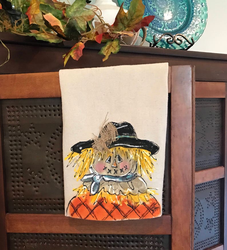 May include: A hand-painted kitchen towel featuring a whimsical scarecrow with a black hat, yellow straw hair, and a blue bandana. The scarecrow is wearing an orange and brown plaid shirt. The towel is made of natural linen and has a rustic, farmhouse style.