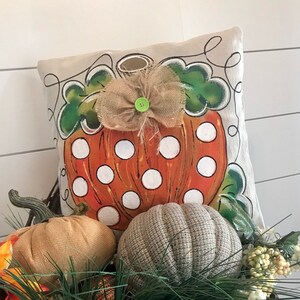 May include: A beige pillow with a painted orange pumpkin with white polka dots and green leaves. A burlap bow with a green button is on top of the pumpkin. The pillow is sitting on a bed of green pine needles and brown and gray pumpkins.