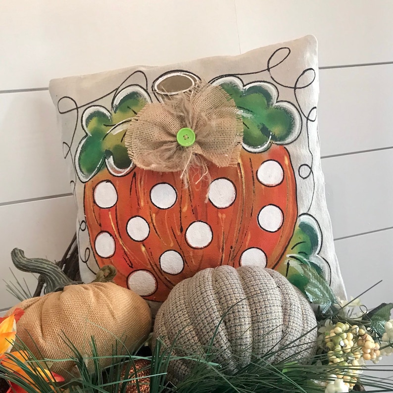 May include: A beige pillow with a painted orange pumpkin with white polka dots and green leaves. A burlap bow with a green button is on top of the pumpkin. The pillow is sitting on a bed of green pine needles and brown and gray pumpkins.