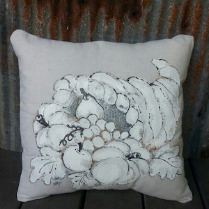 May include: A white throw pillow with a hand-painted design of a cornucopia overflowing with fruit, including grapes, pears, and apples. The pillow is made of a natural linen fabric and has a subtle texture.