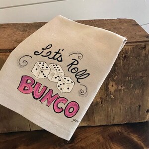 Peut inclure: Une serviette en lin beige avec l'inscription "Let's Roll Bunco" en noir et rose. Trois d&eacute;s blancs &agrave; points noirs sont repr&eacute;sent&eacute;s au centre. La serviette est pli&eacute;e et pos&eacute;e sur une surface en bois.