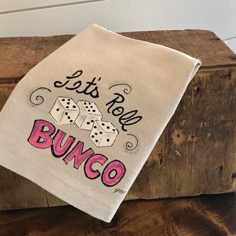 Peut inclure: Une serviette en lin beige avec l'inscription "Let's Roll Bunco" en noir et rose. Trois d&eacute;s blancs &agrave; points noirs sont repr&eacute;sent&eacute;s au centre. La serviette est pli&eacute;e et pos&eacute;e sur une surface en bois.
