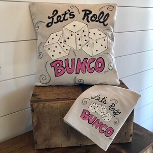 Peut inclure: Un coussin beige et un torchon assorti avec l'inscription "Let's Roll BUNCO" en noir et rose. Le motif comprend trois d&eacute;s blancs avec des points noirs. Les articles sont pr&eacute;sent&eacute;s sur une bo&icirc;te en bois.