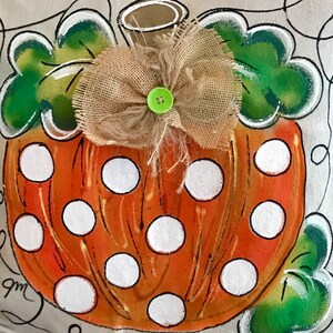 May include: A hand-painted orange pumpkin with white polka dots and a burlap bow with a green button. The pumpkin is surrounded by green leaves.