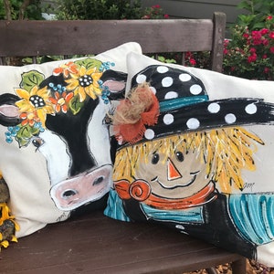 May include: Two decorative throw pillows with hand-painted designs. One pillow features a black and white cow with a floral crown, the other features a scarecrow with a black and white polka dot hat.