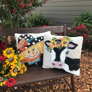 May include: Two decorative throw pillows with a farmhouse theme. One pillow features a black and white cow with a sunflower crown. The other pillow features a scarecrow with a black and white polka dot hat. Both pillows are on a wooden bench.