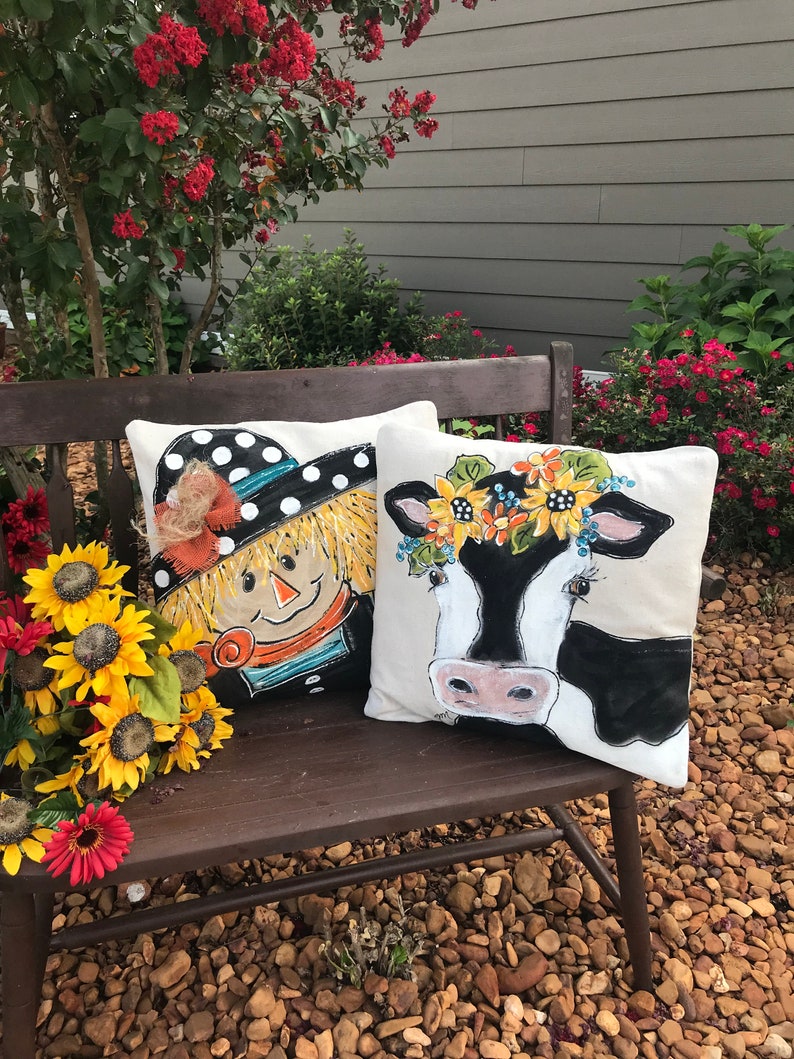 May include: Two decorative throw pillows with a farmhouse theme. One pillow features a black and white cow with a sunflower crown. The other pillow features a scarecrow with a black and white polka dot hat. Both pillows are on a wooden bench.
