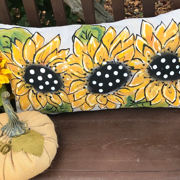 May include: A white pillow with a hand-painted design of three yellow sunflowers with black polka dots on the centers. The sunflowers are on a white background with green leaves.